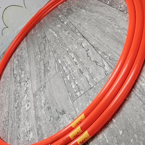 Puede incluir: Tres aros de hula-hoop de color naranja brillante, cada uno con una etiqueta amarilla que indica una medida de 76 cm. Los aros están dispuestos en una configuración anidada, mostrando su forma circular y su color vibrante. El fondo es una superficie gris texturizada.