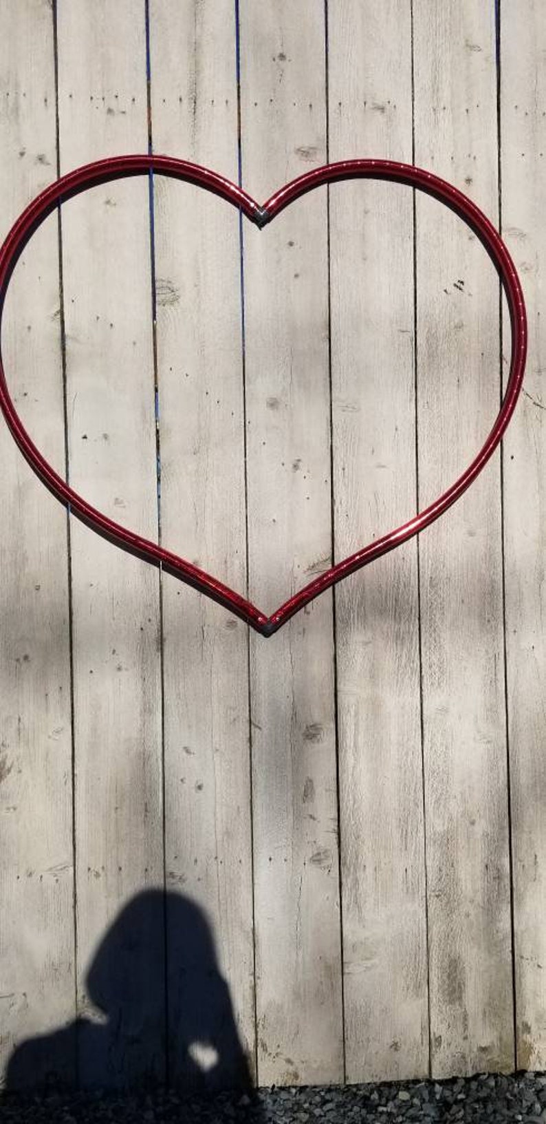 May include: A red heart-shaped metal hoop is attached to a white wooden fence. The hoop is made of thin metal and is slightly bent in the middle. The fence is made of vertical wooden planks.