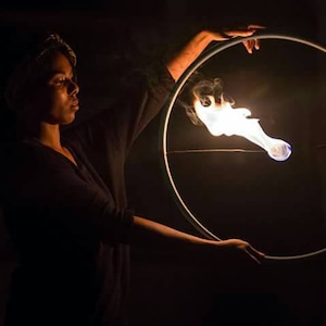 May include: A person holds a glowing ring with a flame in the center. The flame emits white smoke against a dark background. The ring is held up with both hands, creating a circular shape.