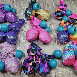 May include: A collection of colourful fabric-covered items with teal spherical accents. The fabrics are various shades of purple, pink, gold, and patterned designs. The items are arranged on a wooden surface.