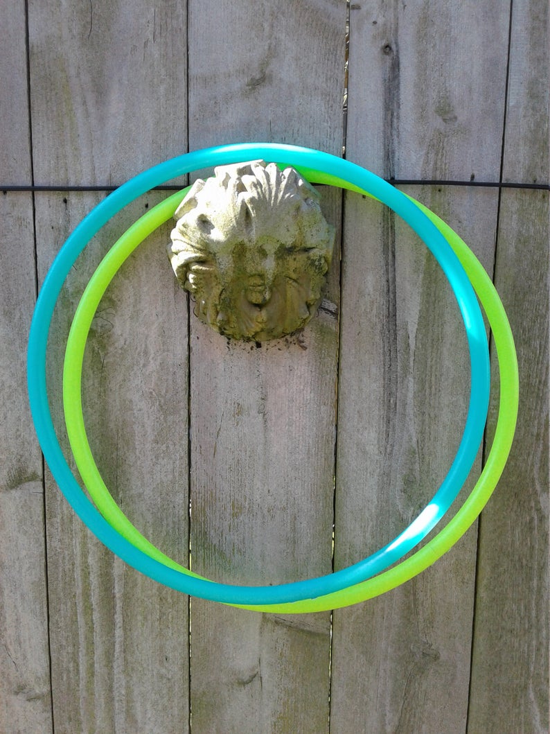 May include: Two plastic hoops, one green and one blue, are intertwined and hanging on a wooden fence. The hoops are both circular and appear to be made of flexible plastic.