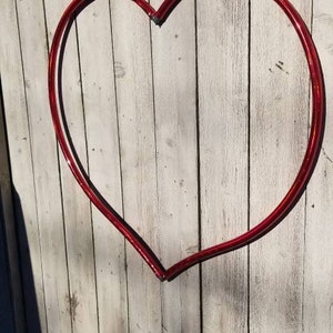 May include: A red heart-shaped metal hoop is attached to a white wooden fence. The hoop is made of a single piece of metal that is bent into a heart shape.