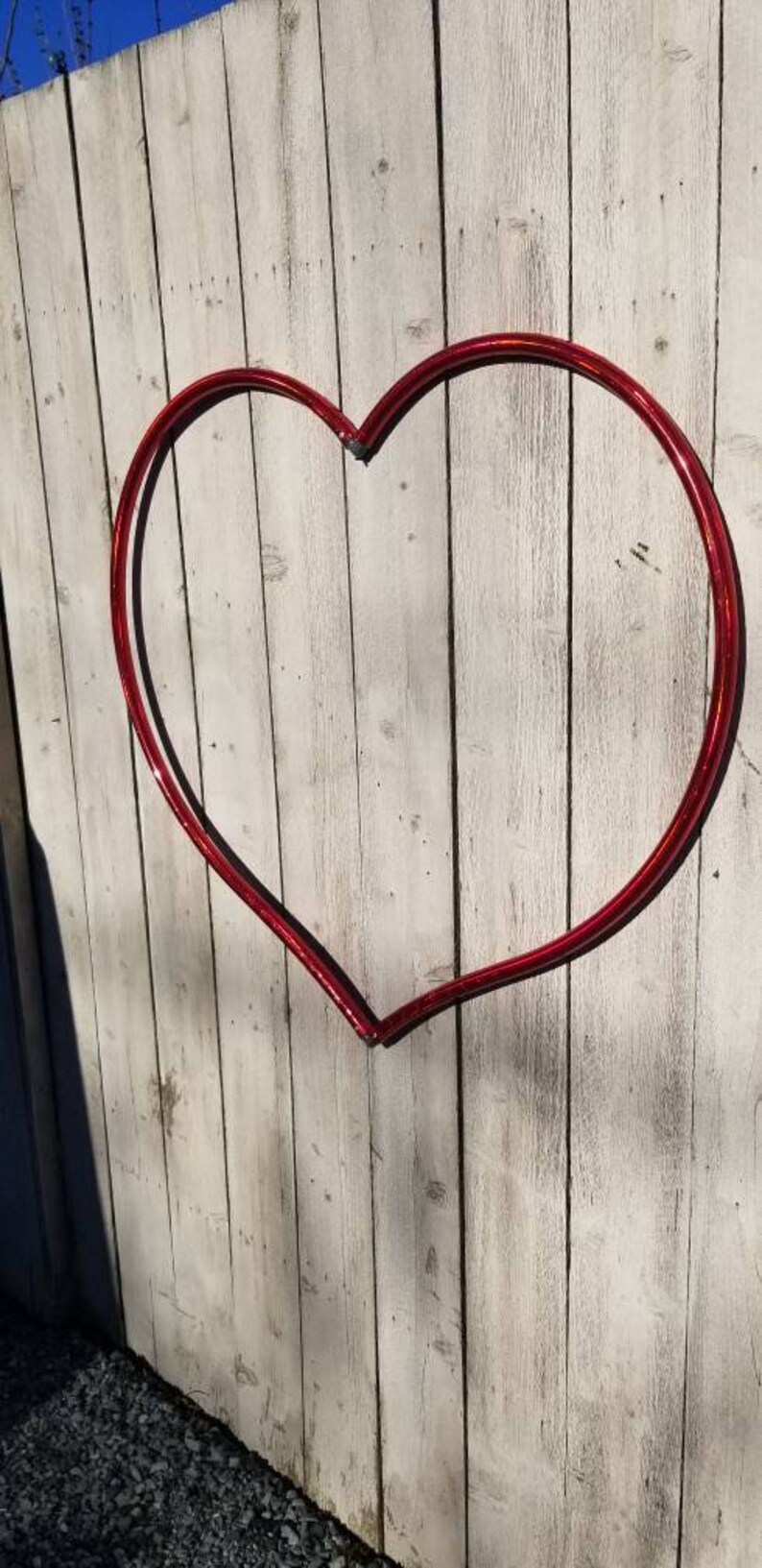 May include: A red heart-shaped metal hoop is attached to a white wooden fence. The hoop is made of a single piece of metal that is bent into a heart shape.