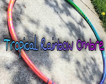 Könnte beinhalten: Ein bunter Regenbogen-Ombre-Hula-Hoop mit rosa, orange, gelben, grünen, blauen und violetten Streifen. Der Reifen liegt auf einer grauen Betonfläche. "Tropical Rainbow Ombre" ist in blauem und violettem Text auf der Oberfläche geschrieben.