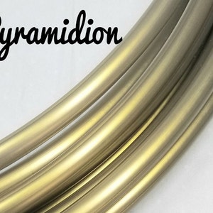 May include: Close-up of several curved, metallic tubes with a gold and bronze finish. The word "Pyramidion" is written in a cursive font in the upper left corner. The tubes are arranged in a flowing, parallel pattern against a white background.