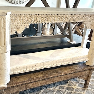 May include: Whitewashed wood console table with intricate carvings and a distressed finish. The table has a rectangular top and two ornate columns.