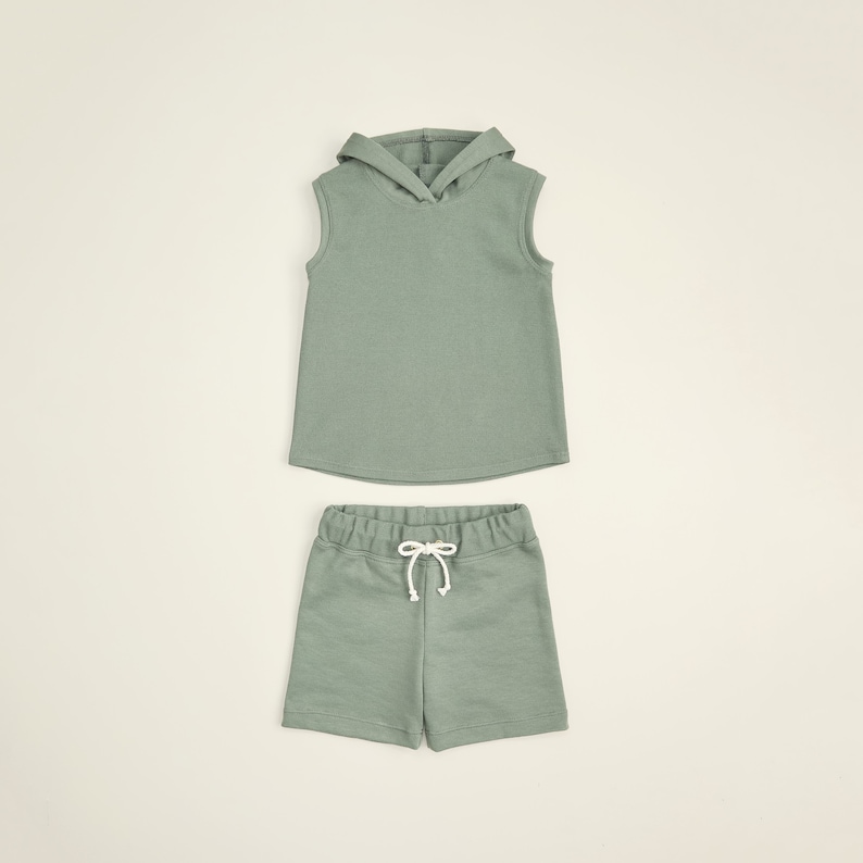 May include: A sage green sleeveless hoodie and matching shorts set for kids. The hoodie has a drawstring hood and the shorts have a drawstring waist.