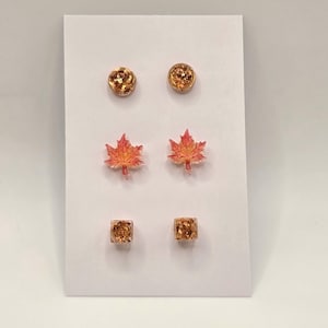 May include: A set of three pairs of stud earrings displayed on a white card. The top pair are round with gold flakes, the middle pair are shaped like maple leaves in shades of orange and pink, and the bottom pair are square with gold flakes.