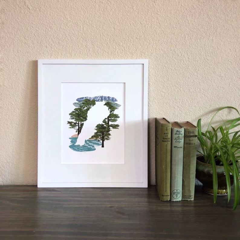 May include: A white framed print of a silhouette of a bird with a mountain and trees inside the silhouette. The print is on a wooden table with three books and a potted plant.