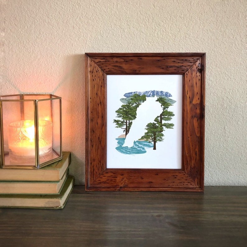 May include: A framed print of a white silhouette of a mountain range with a blue and green landscape behind it. The frame is made of dark wood.