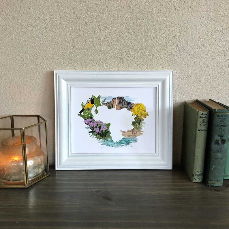 May include: A white framed print of a yellow bird perched on a branch with a mountain range in the background. The print is surrounded by a border of green leaves and purple flowers.