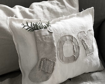 Puede incluir: Almohada blanca con la palabra "JOY" cosida en ella en un estilo rústico. La almohada tiene un bolsillo con una ramita de vegetación que sobresale.