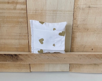 Puede incluir: Pequeña bolsa de tela blanca con cierre de solapa. La bolsa está decorada con formas de corazón doradas. El fondo es una superficie de madera.