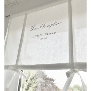 Könnte beinhalten: Weißer Leinen-Fensterrollo mit dem aufgedruckten Text "The Hamptons Long Island New York". Der Rollo ist mit weißem Band zurück gebunden.