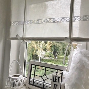 May include: White sheer curtains with a delicate lace trim hang in a window. The curtains are tied back with white ribbon. A white wicker lantern, a framed windowpane, and a wooden sign with the text "Let it be." are on the windowsill.