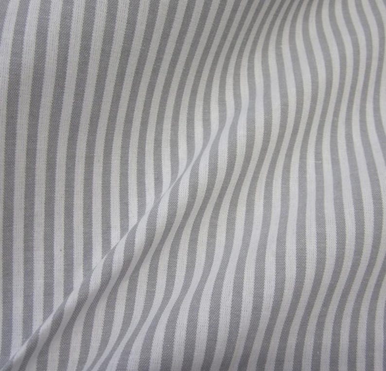 Puede incluir: Una pieza de tela con un patr&oacute;n repetitivo de rayas verticales en tonos alternos de gris claro y blanco. El tejido muestra pliegues y drapeados suaves, creando un efecto de onda sutil a trav&eacute;s de la superficie del material.