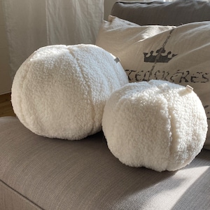 Könnte beinhalten: Zwei weiße, flauschige runde Kissen auf einem grauen Sofa. Die Kissen sind aus einem weichen, flauschigen Material gefertigt und haben eine leicht strukturierte Oberfläche.