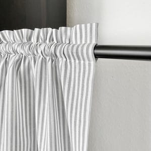 May include: A close-up shot of a grey and white striped curtain panel. The fabric is gathered at the top and suspended from a black curtain rod. The vertical stripes are evenly spaced, creating a classic look. The curtain is against a white wall.