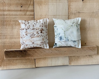 May include: Two small, rectangular pillows with floral patterns. The pillows are sitting on a wooden shelf. The pillow on the left has a brown and white floral pattern. The pillow on the right has a blue and white floral pattern.
