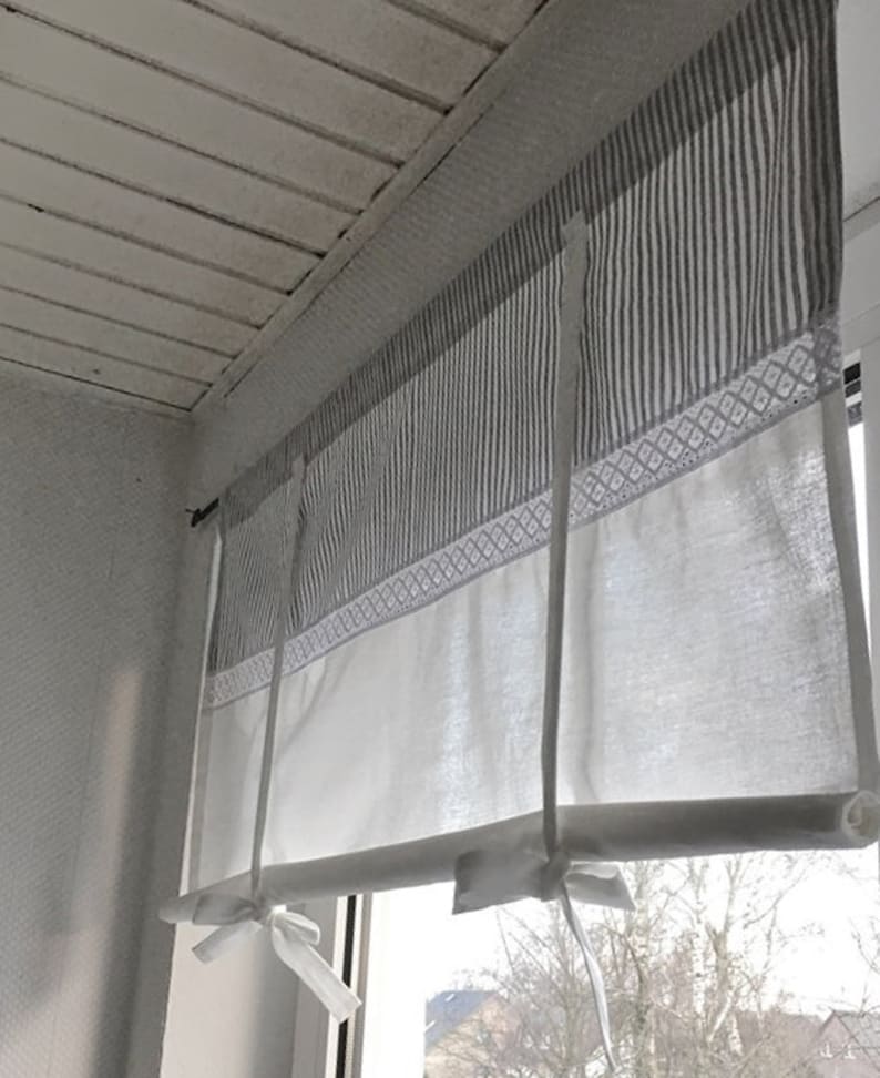Puede incluir: Una cortina de ventana con lazos que presenta una secci&oacute;n superior con finas rayas verticales grises y blancas, una banda central de bordado geom&eacute;trico blanco y una secci&oacute;n inferior blanca s&oacute;lida que se enrolla y asegura con dos lazos.
