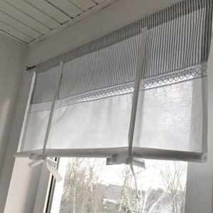 Könnte beinhalten: Ein weißer und grauer gestreifter Stoff-Fensterüberhang mit weißer Spitzenborte und weißen Stoffbändern. Der Überhang ist an einem weißen Fensterrahmen befestigt.