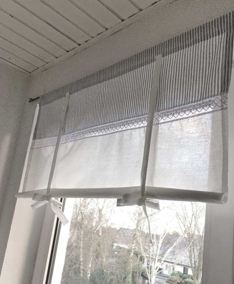 Puede incluir: Una cortina de ventana que presenta una secci&oacute;n superior con rayas verticales grises y blancas, una banda horizontal de encaje blanco y una secci&oacute;n inferior blanca s&oacute;lida. La cortina se mantiene enrollada mediante dos lazos de tela blanca.