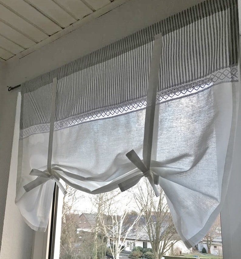 Puede incluir: Una cortina blanca de atar con un panel superior de rayas verticales grises y blancas. La cortina incluye una banda decorativa de encaje blanco que separa la secci&oacute;n rayada de la parte inferior blanca lisa, recogida mediante dos lazos de tela.