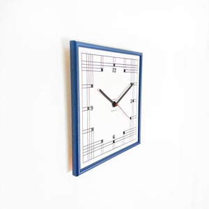 May include: A square clock with a white face and black Roman numeral hour markers. The clock has a blue frame and a geometric design on the face. The clock hands are black and red.