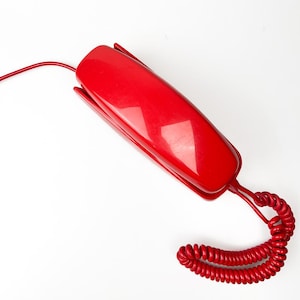 May include: A bright red vintage telephone receiver with a matching coiled cord. The receiver has a sleek, rectangular design with a glossy finish. The cord extends to the left and curls at the bottom, suggesting a classic, retro aesthetic.