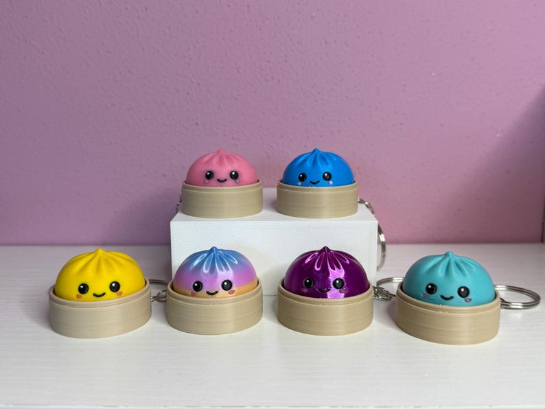 Puede incluir: Seis peque&ntilde;os llaveros redondos y coloridos con forma de dumpling, cada uno sobre una cesta de vapor beige en miniatura. Los dumplings tienen caras sonrientes con ojos negros y mejillas rosadas, en rosa, azul, amarillo, degradado violeta-rosa, violeta met&aacute;lico y verde azulado.