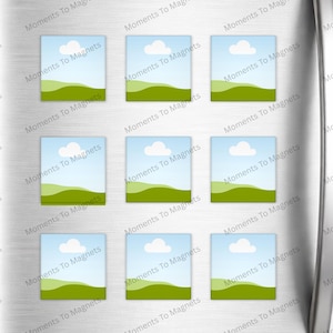 May include: Nine rectangular magnets with a landscape design are displayed on a silver refrigerator door. Each magnet features a blue sky with a white cloud, green hills, and the text "Moments To Magnets".