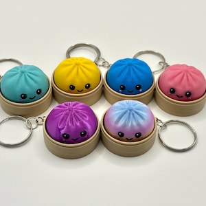 Puede incluir: Seis llaveros con figuras redondas y coloridas de dumplings con caras sonrientes, cada uno dentro de un peque&ntilde;o recipiente cil&iacute;ndrico de color tostado. Los dumplings son de color verde azulado, amarillo, azul, rosa, morado y degradado.