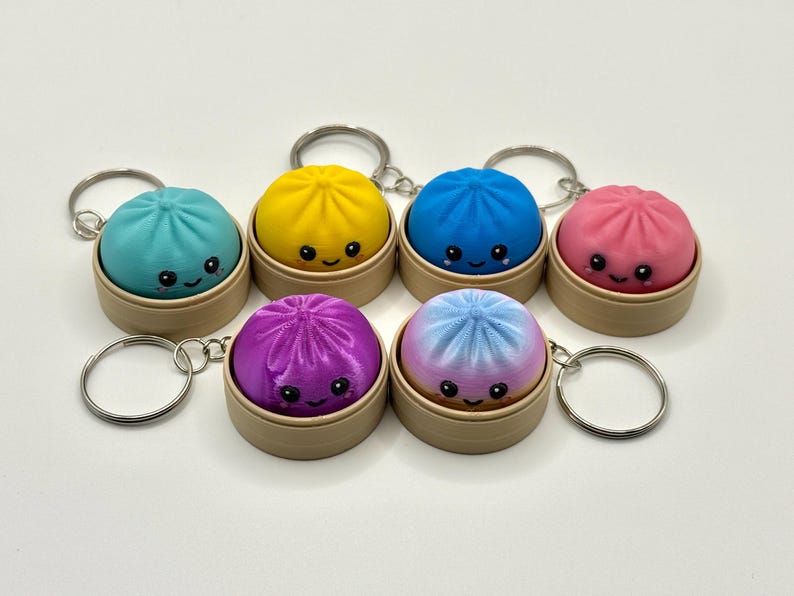 Puede incluir: Seis llaveros con figuras redondas y coloridas de dumplings con caras sonrientes, cada uno dentro de un peque&ntilde;o recipiente cil&iacute;ndrico de color tostado. Los dumplings son de color verde azulado, amarillo, azul, rosa, morado y degradado.