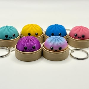 Puede incluir: Una colecci&oacute;n de seis peque&ntilde;os llaveros redondos y coloridos con forma de bola de masa, cada uno en una cesta de vapor en miniatura de color tostado. Las bolas tienen caras sonrientes con ojos negros y mejillas rosadas, en colores turquesa, amarillo, azul, rosa, morado y degradado.