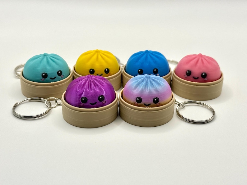 Puede incluir: Una colecci&oacute;n de seis peque&ntilde;os llaveros redondos y coloridos con forma de bola de masa, cada uno en una cesta de vapor en miniatura de color tostado. Las bolas tienen caras sonrientes con ojos negros y mejillas rosadas, en colores turquesa, amarillo, azul, rosa, morado y degradado.