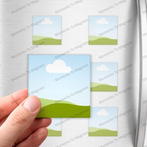 May include: A hand holding a square magnet with a landscape design of a blue sky, white cloud, and green hills. Several similar magnets are attached to a refrigerator. The text "Moments To Magnets" is printed on the magnets.