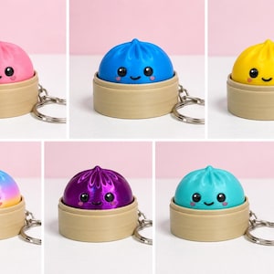 Puede incluir: Una colecci&oacute;n de seis llaveros de dumpling, cada uno con un personaje colorido y sonriente dentro de una base de vaporera circular de color tostado. Los dumplings vienen en rosa, azul, amarillo, turquesa, un acabado met&aacute;lico morado y degradado.