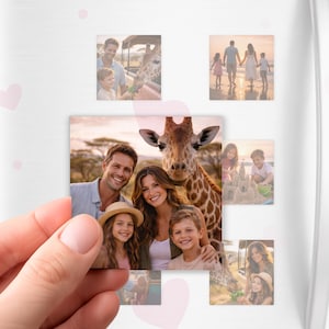 May include: A hand holds a rectangular photo magnet featuring a family with a giraffe. Other photo magnets on a white surface show family photos, including a beach scene and a child feeding a giraffe. Pink heart stickers are scattered around.