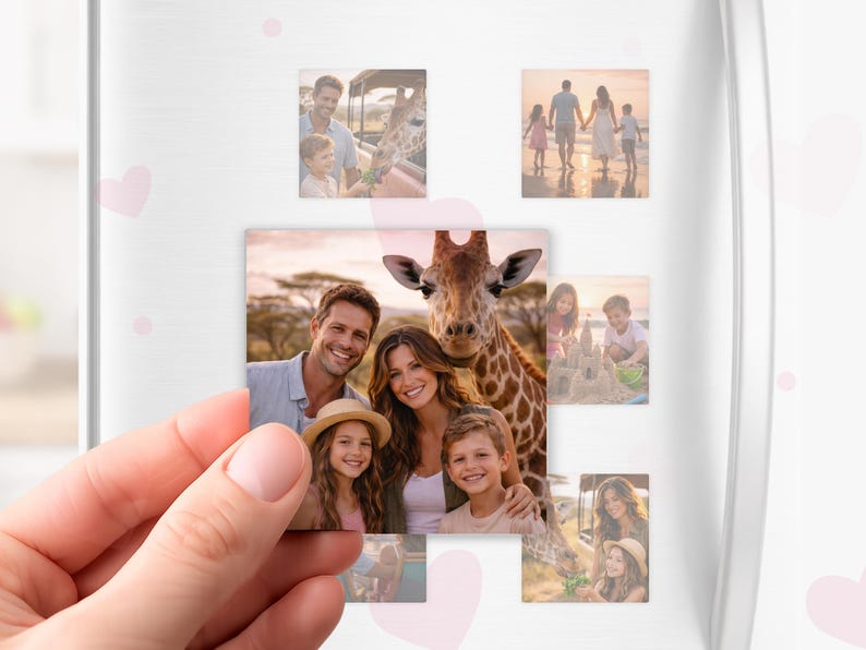 May include: A hand holds a rectangular photo magnet featuring a family with a giraffe. Other photo magnets on a white surface show family photos, including a beach scene and a child feeding a giraffe. Pink heart stickers are scattered around.