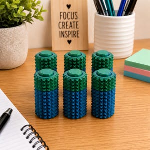 May include: Six cylindrical sensory fidget tools arranged in two rows of three. Each tool features a textured surface with small, raised pyramid-shaped spikes. The top half of each cylinder is green, while the bottom half is blue, with a flat top.