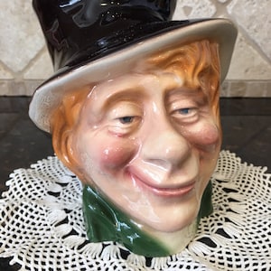 May include: A ceramic novelty mug shaped like a smiling man's head wearing a black top hat. The mug features a detailed face with red cheeks, orange hair, and a green collar. The mug is sitting on a white lace doily.