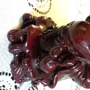 May include: A red, smiling, chubby figurine of a Buddha sitting on a base. The Buddha is wearing a robe and holding a bag in one hand.