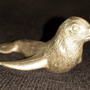 May include: A silver figurine of a seal, lying on its belly with its flippers outstretched. The seal has a detailed, textured surface and realistic features.