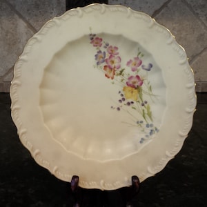 May include: A white porcelain plate with a gold rim and a floral design. The flowers are pink, purple, yellow, and blue.