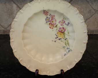 May include: A white porcelain plate with a gold rim and a floral design. The flowers are pink, purple, yellow, and blue.