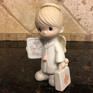 May include: A porcelain figurine of an angel wearing a white doctor's coat and a halo. The angel is holding a sign that says "it's a Perfect Boy" and a small medical bag with a red cross.