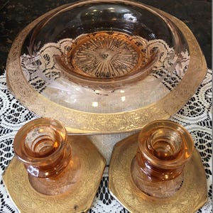 May include: A set of three vintage glass pieces in a pink hue with gold accents. The set includes a large bowl and two candle holders. The bowl has a scalloped edge and a decorative pattern on the bottom. The candle holders have a simple design and are perfect for adding a touch of elegance to any table setting.