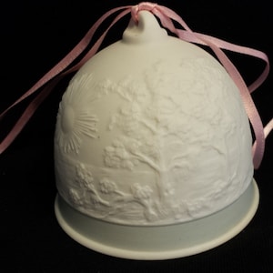May include: A white porcelain bell ornament with a raised design of a tree and a sun. The bell has a light blue base and is tied with a pink ribbon.