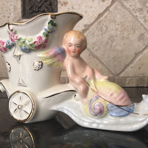 May include: A white ceramic vase shaped like a chariot pulled by a cherub riding a seahorse. The vase is decorated with gold trim, pink and blue flowers, and a cherub with wings.