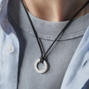 May include: A silver necklace with a round pendant featuring Roman numerals. The necklace is on a black cord and is worn by a person wearing a light blue shirt.